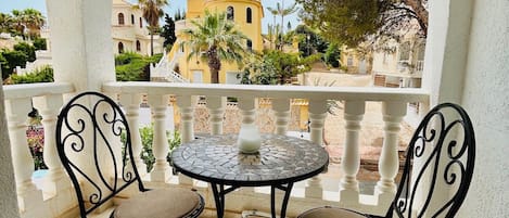 En balkon med to stole og et bord, der udover et hus med gul facade og et træ.