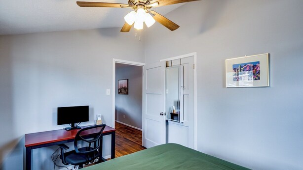 A bedroom with a bed, a desk with a computer, a chair, a ceiling fan, and a door.