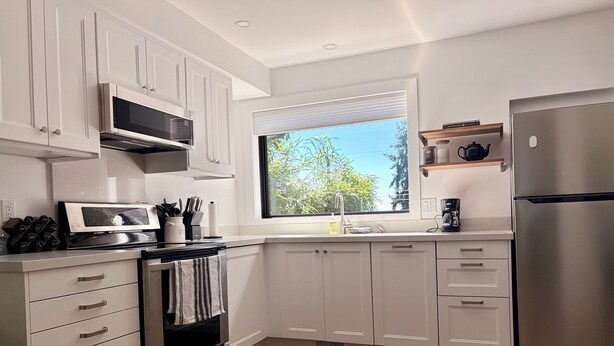 A modern kitchen with white cabinetry, a stainless steel refrigerator, and a window providing a view of greenery outside. - Modern and Bright Lynn Valley Retreat (North Vancouver)