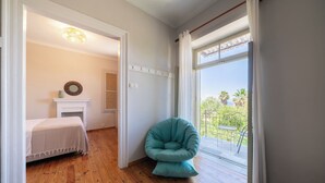 Una habitación con piso de madera, una cama con cubrecama blanco, una silla color verde azulado, una chimenea y vistas a un balcón con plantas.