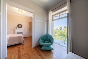 A room with a wooden floor, a bed with a white bedspread, a teal chair, a fireplace, and a view of a balcony with plants. - Meraki Villa Pelion Home to Home Management (Kalamos Magnesia)