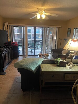 A living room with a television, a table with decorative items, a ceiling fan, and a view of a balcony with outdoor furniture.