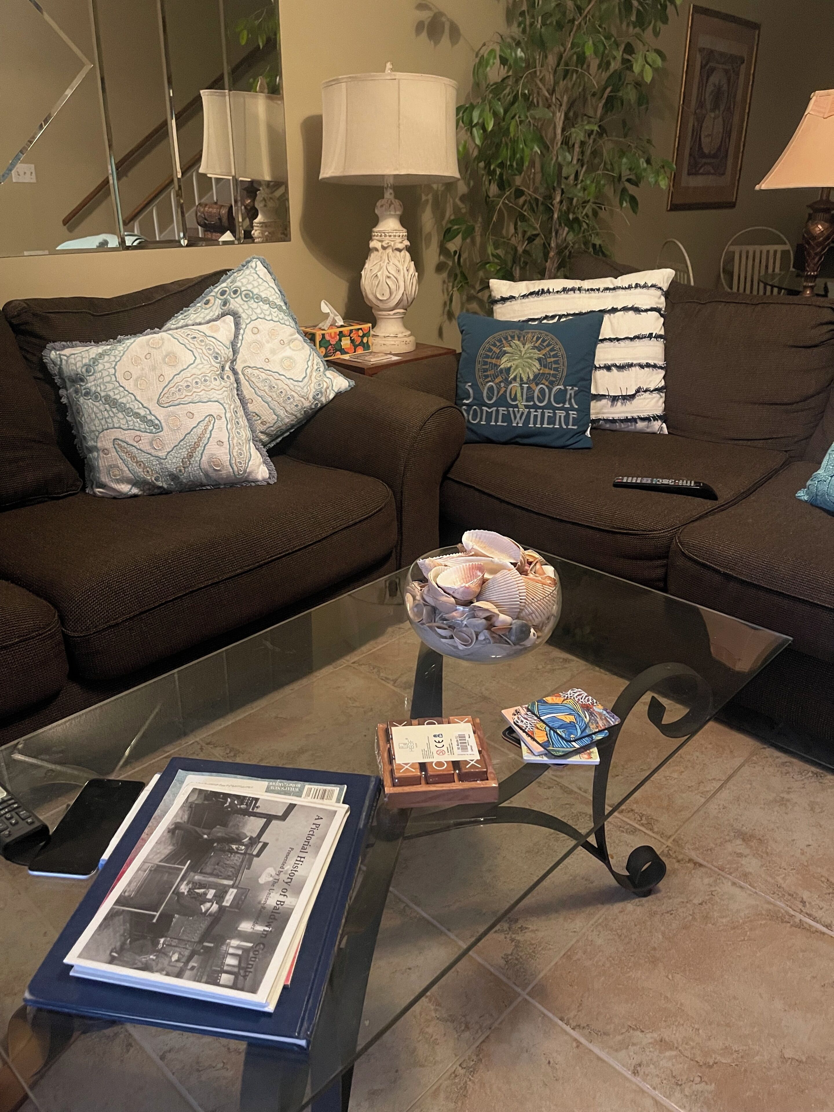 A living room with a glass coffee table, a sofa with patterned cushions, a lamp, and a framed picture on the wall.
