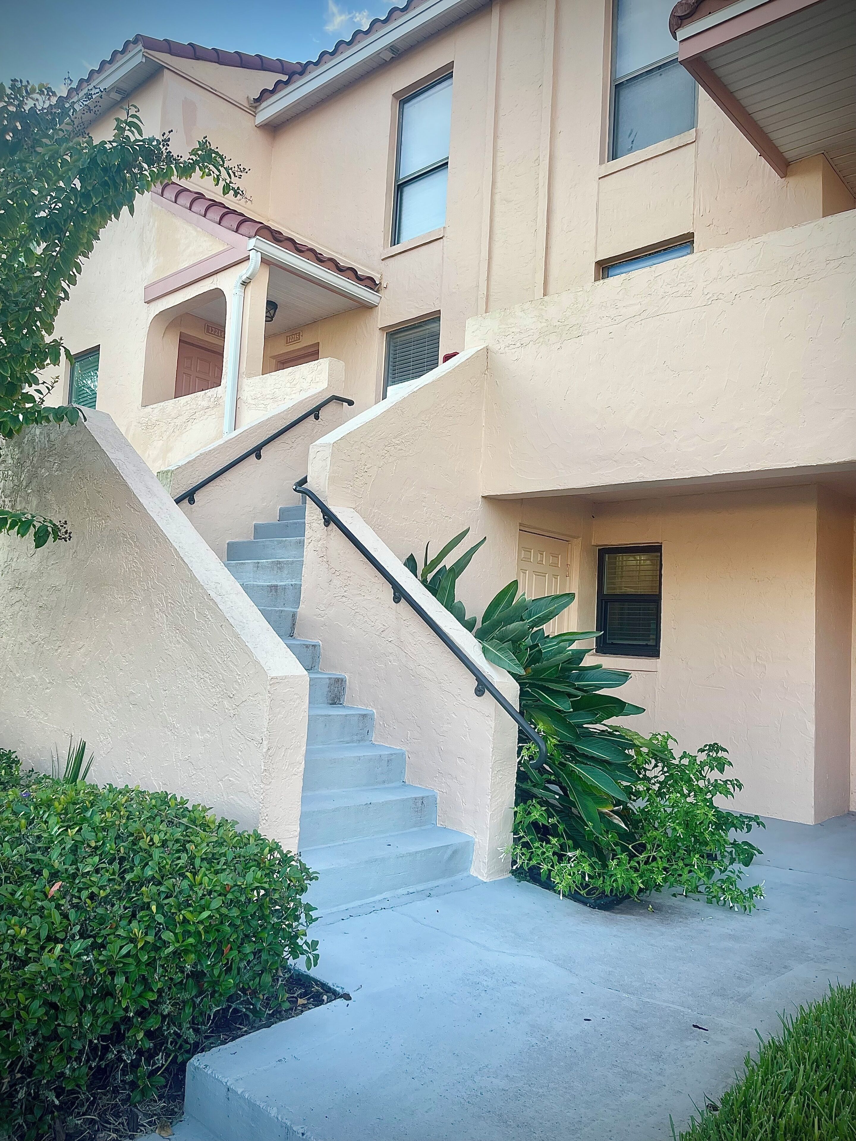Un edificio residencial con escaleras de hormigón, varias ventanas y abundante vegetación alrededor.