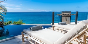 Un salon de jardin aménagé au bord de la piscine, avec deux chaises longues, un barbecue et une vue sur l’océan.