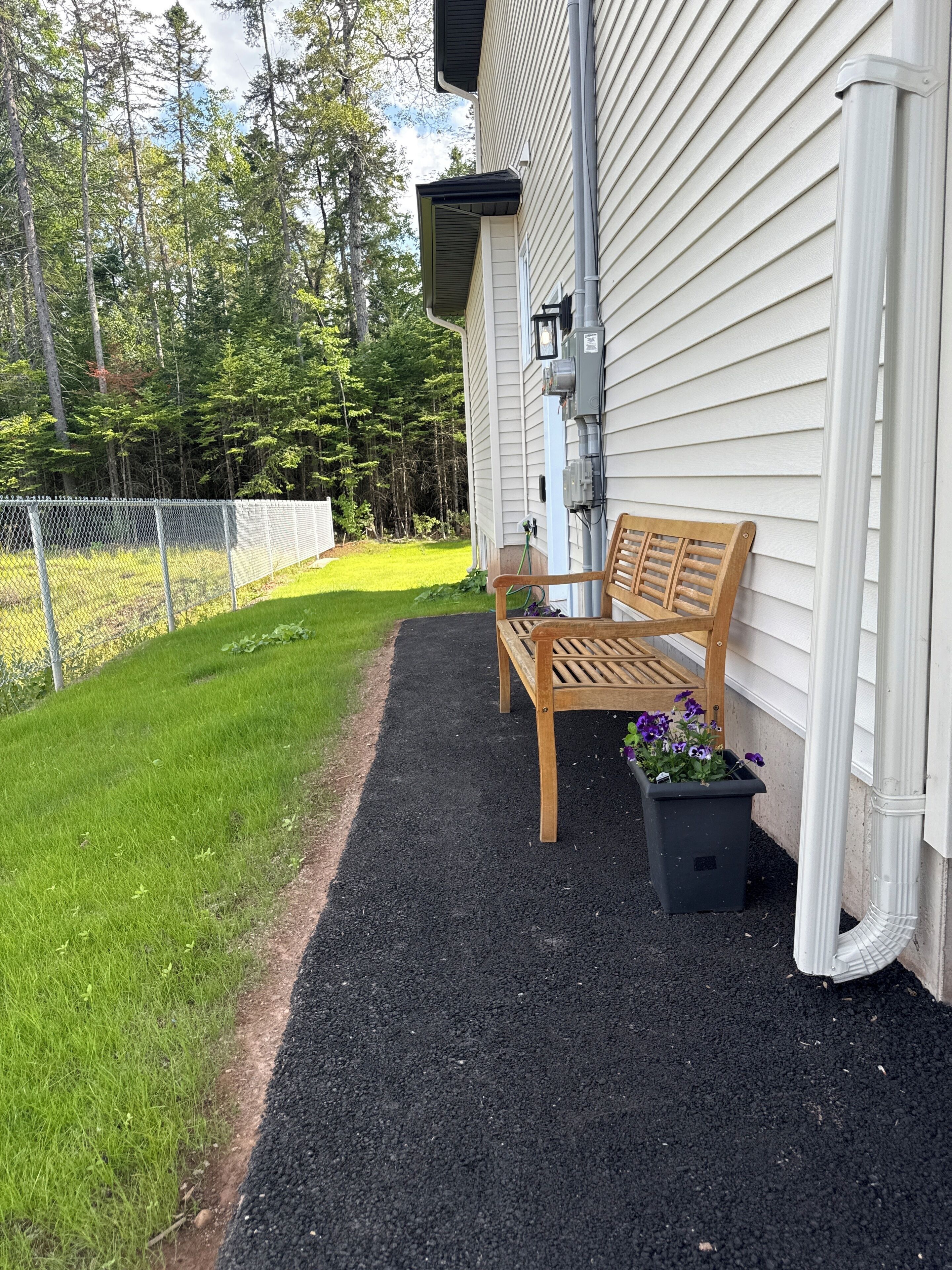 A wooden bench outside a house with a black ground cover and a flower pot with purple flowers.