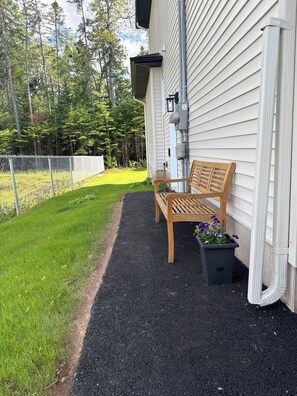 A wooden bench outside a house with a black ground cover and a flower pot with purple flowers.
