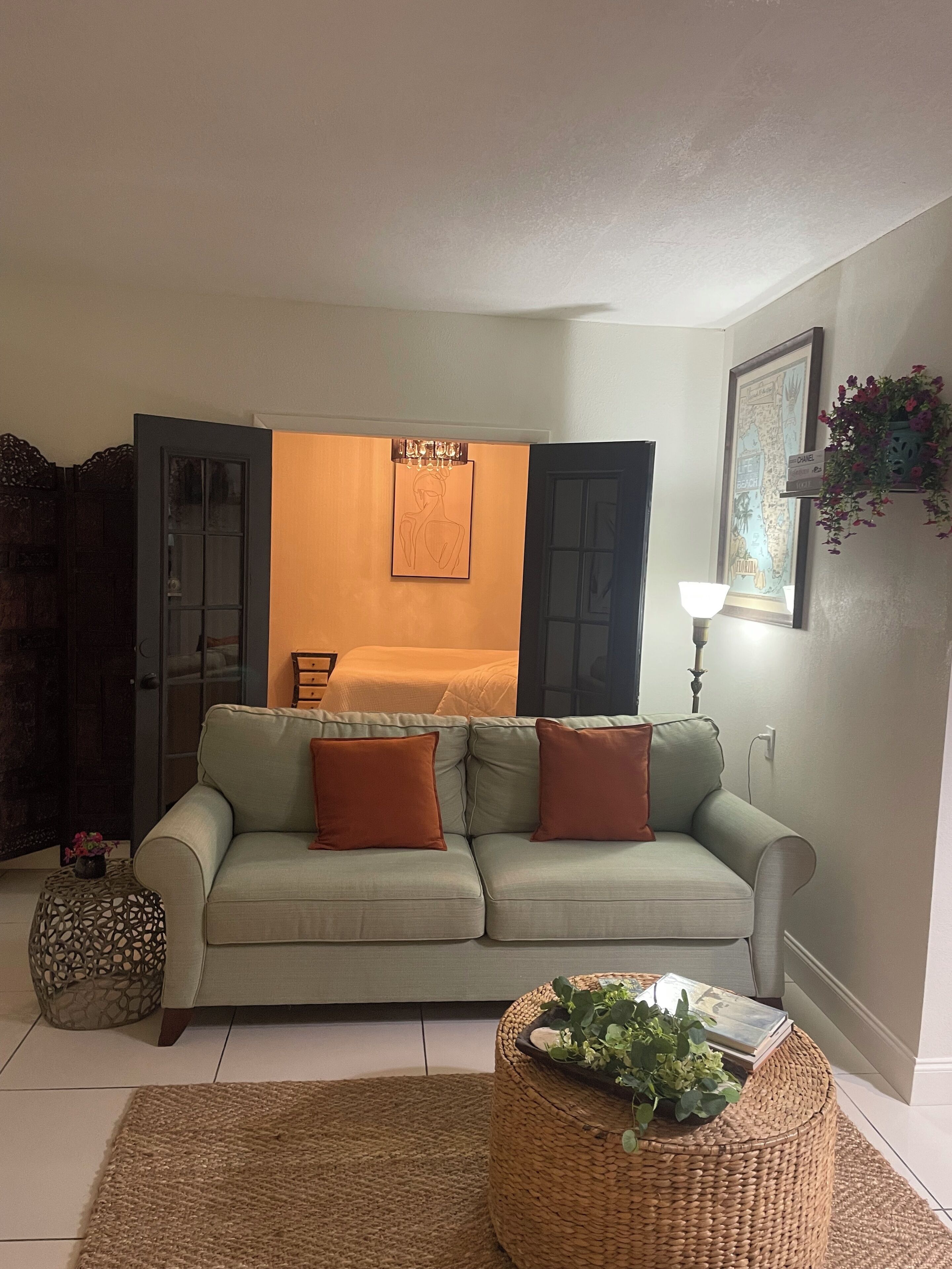 A living room with a sofa, a small round table with a plant, a wall-mounted lamp, and a door leading to another room.