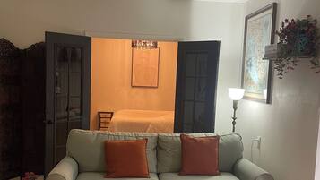 A living room with a sofa, a small round table with a plant, a wall-mounted lamp, and a door leading to another room.