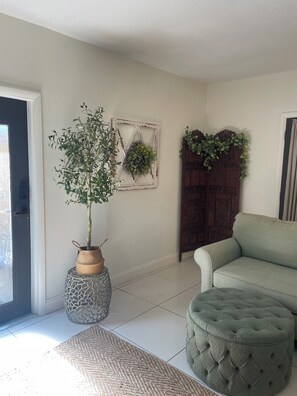 A room with a green sofa, a round ottoman, a potted plant, a decorative wall piece, and a wooden door with greenery. - Lovely “Dreamy Nook at Cocoa Village” (Cocoa)