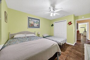 A bedroom with two beds, a ceiling fan, a door with a decorative wreath, and a hallway leading to another room. - Steps To Beach Access 45 & Salty Goat Saloon (Panama City Beach)