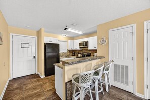 A kitchen with white cabinets, a black refrigerator, a granite countertop, and a bar area with white chairs. - Steps To Beach Access 45 & Salty Goat Saloon (Panama City Beach)