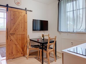 Una habitación con puerta corredera de madera, televisor montado en la pared, mesa de comedor con sillas y ventana con cortinas.