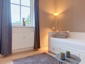 A bedroom with a bed, a bedside lamp, a window with curtains, a radiator, a small table with plants, and a wall-mounted shelf.