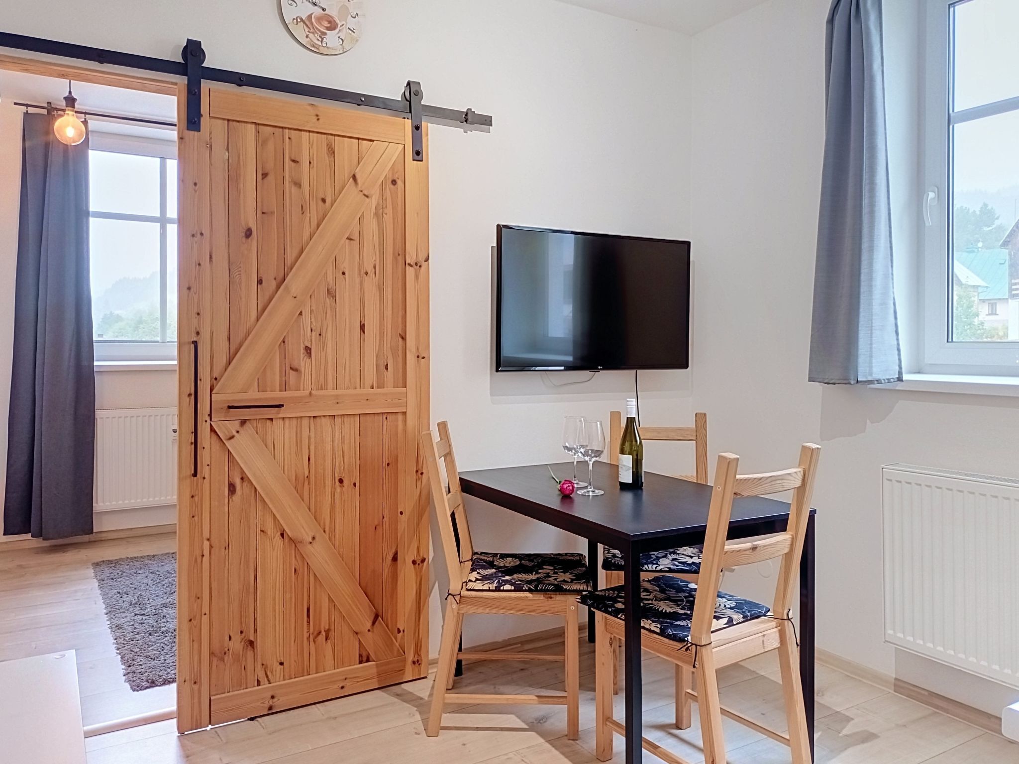 A dining area with a wooden table, four chairs, a TV mounted on the wall, and a sliding barn door.
