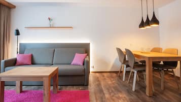 A modern living room with a grey sofa, a wooden coffee table, a dining table with chairs, and a pink carpet.