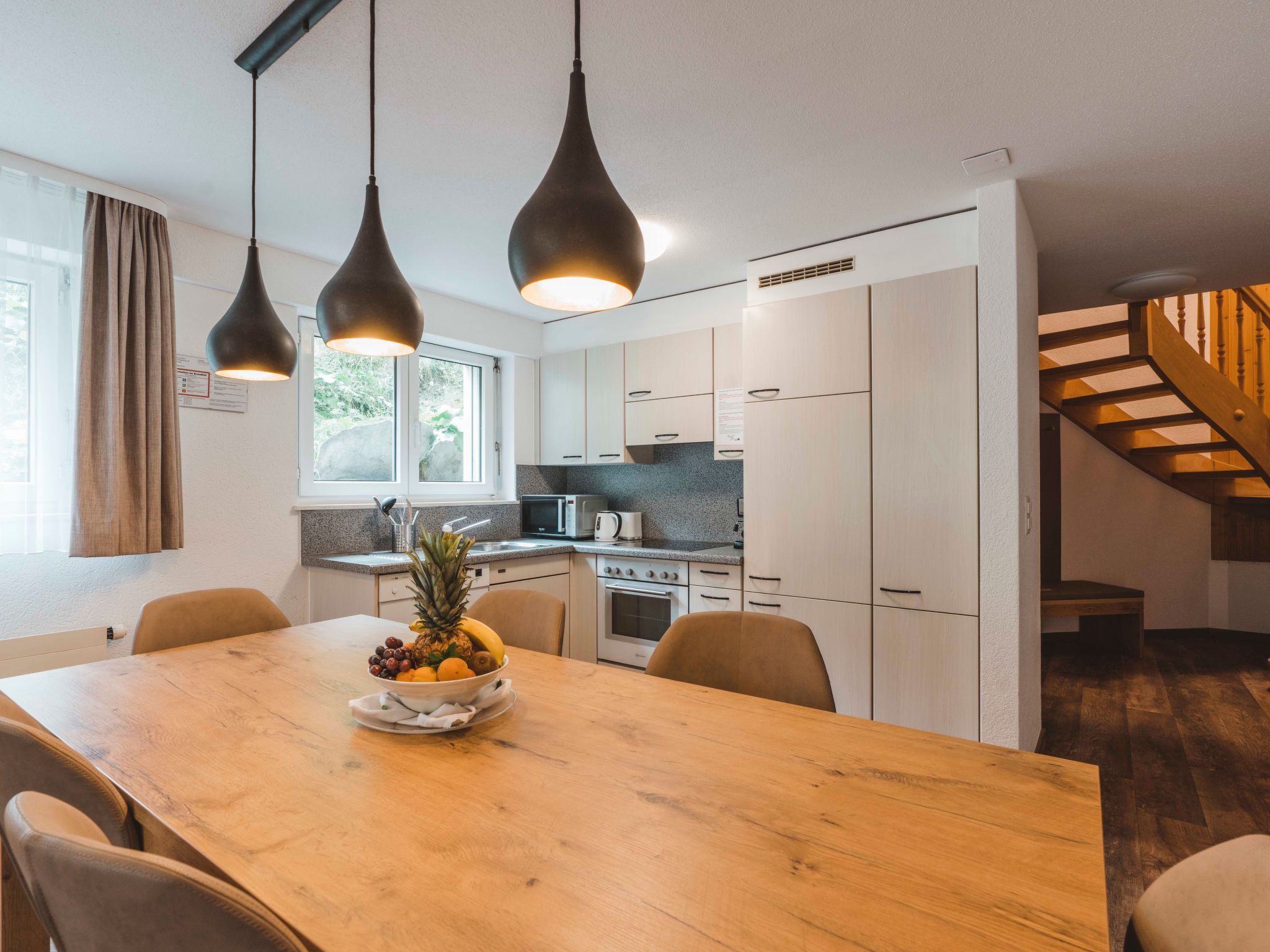 A modern kitchen with a wooden dining table, overhead pendant lights, and a view of the outdoors through a window.
