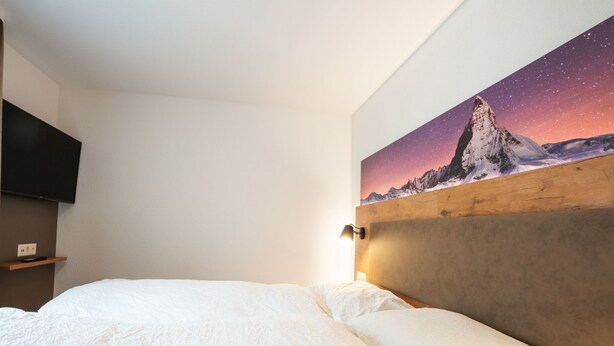Un dormitorio con cama, televisor montado y un cuadro de un paisaje montañoso.
