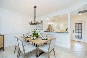 A dining area with a wooden table set for four, surrounded by chairs, a modern chandelier, and a kitchen visible in the background. - Newly Remodeled Naples Retreat - Resort-Style Living at Bridgewater Bay (Naples)