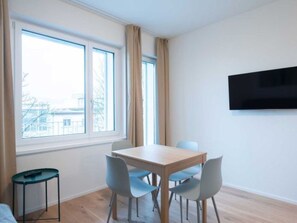 A modern room with a wooden table and four chairs, a small side table, a mounted TV, and a window with curtains. - ZH Green 3 - Letzigrund HITrental Apartment by Interhome (Zurich)