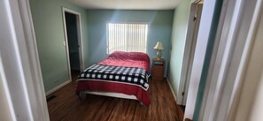 Una habitación estrecha con una cama, una ventana con persianas, una mesita de noche con lámpara y una puerta.