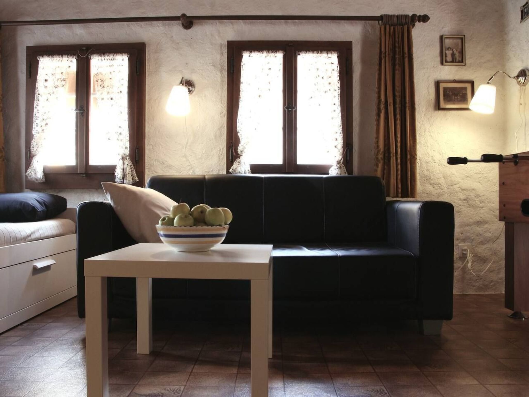 A living room with a sofa, a small table with a bowl of fruit, and two windows with curtains.