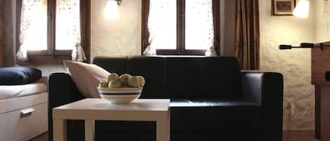 A living room with a sofa, a small table with a bowl of fruit, and two windows with curtains.