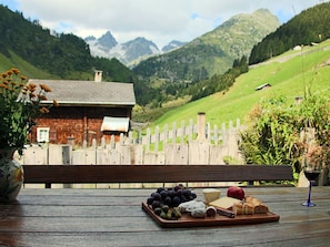 A wooden table with a cheese platter, grapes, a red apple, and a wine glass. A wooden fence and a house with a chimney are visible in the background. - Gabrielenhaus, (Wassen UR), by Interhome (Wassen UR)