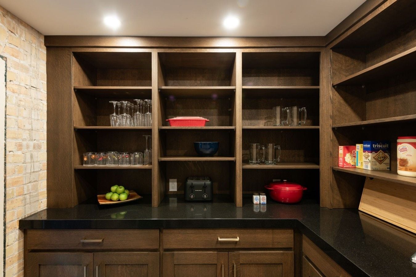 Una cucina con mobili in legno, un piano di lavoro nero e diversi oggetti da cucina, tra cui una pentola rossa, una macchina da caffè e una ciotola di mele.