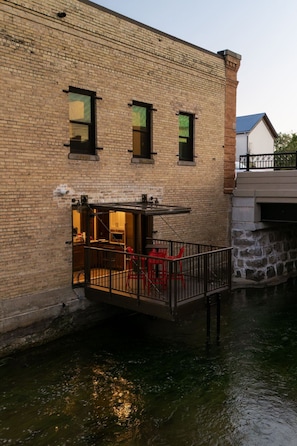 Un bâtiment en briques doté d’un petit espace extérieur avec des chaises rouges et une table, donnant sur une étendue d’eau.