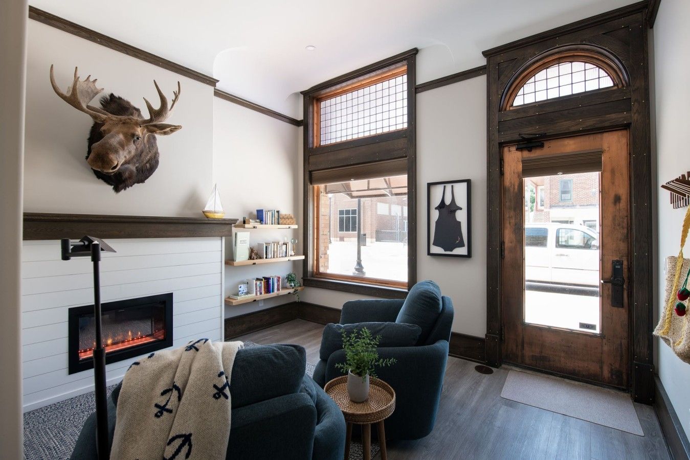 A cozy living room with a fireplace, a mounted moose head, a bookshelf, and a large window.
