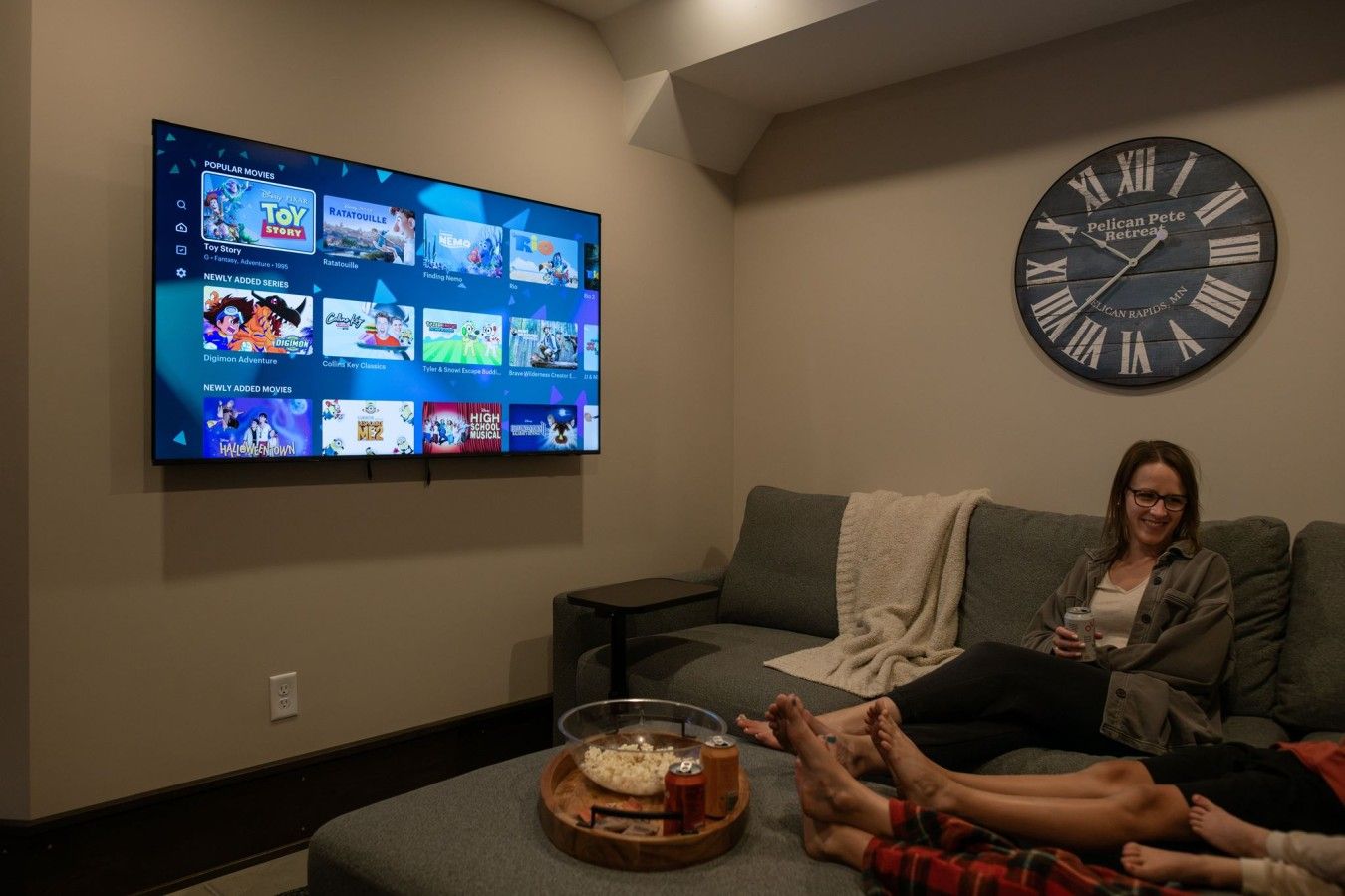 A living room with a flat-screen TV displaying a streaming service interface, a wall clock, and a cozy seating area with a blanket and snacks.