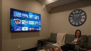 En stue med et fladskærms-tv, der viser en streamingtjenestes brugerflade, et vægur og en hyggelig siddekrog med et tæppe og lidt snacks.