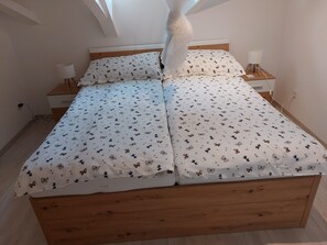 A bedroom with a wooden bed, white bedding with a butterfly pattern, a bedside table with a lamp, and a power outlet on the wall.