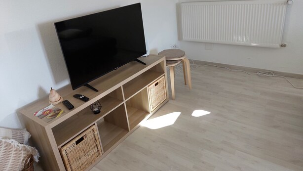 A room with a large flat-screen TV mounted on a wooden console, a radiator on the wall, and a wicker basket under the console. - Les Gîtes du Gore Virat, (Corcelles BE), by Interhome (Corcelles)