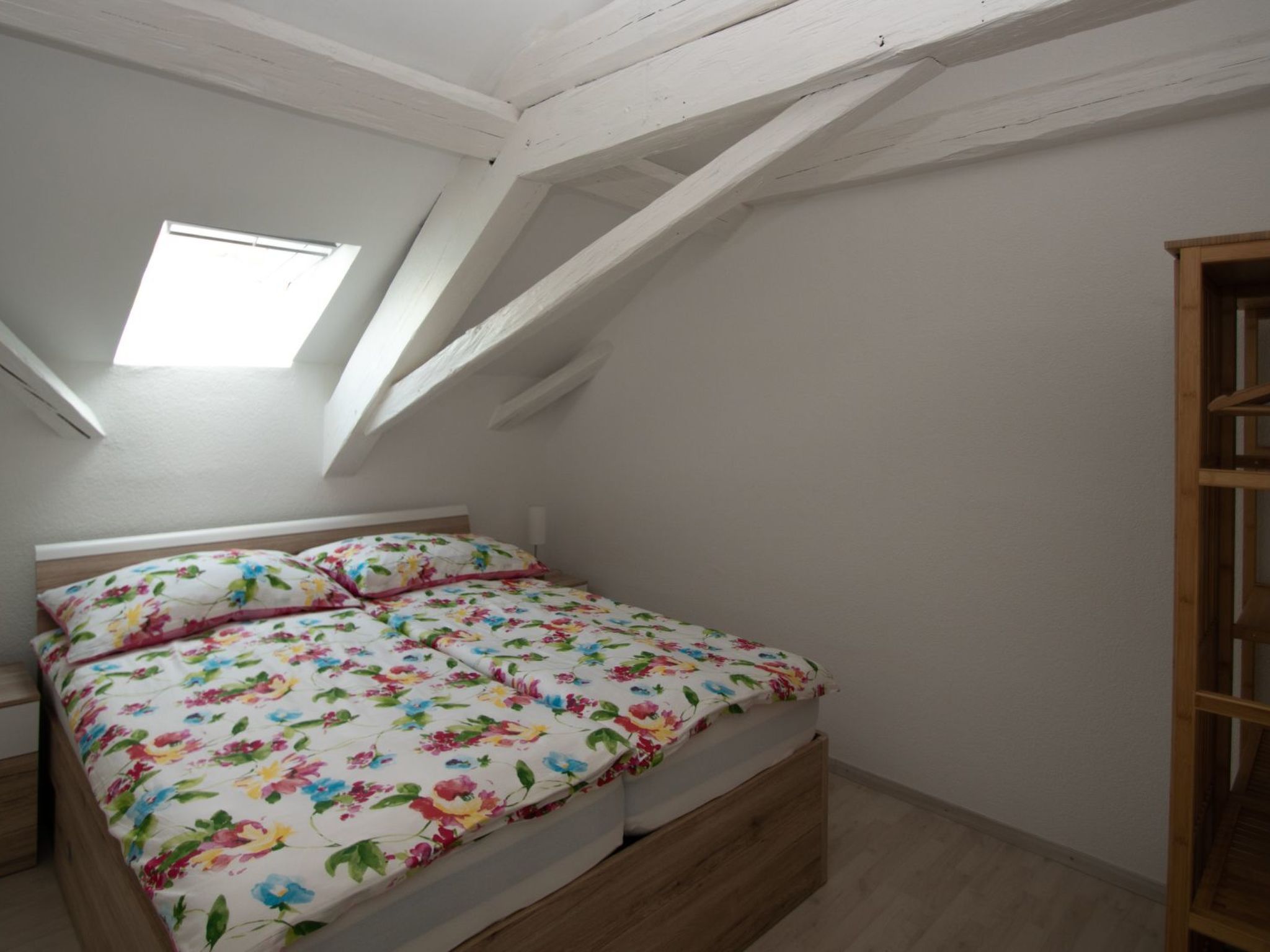 Una camera da letto con soffitto spiovente, un letto con lenzuola a motivi floreali, un comodino e un armadio in legno.