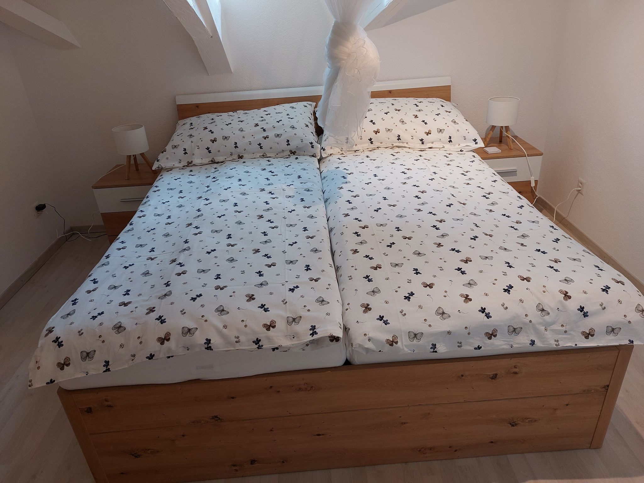 Ein Schlafzimmer mit einem hölzernen Bett, weißer Bettwäsche mit Schmetterlingsmuster, einem Nachttisch mit Lampe und einer Steckdose an der Wand.