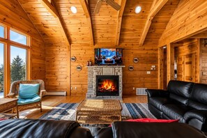 Interior de una cabaña de madera con chimenea de piedra, un televisor, muebles de mimbre y un sofá de cuero.