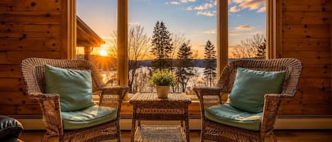A room with two wicker chairs, a small table, and a view of a sunset over a forested landscape.