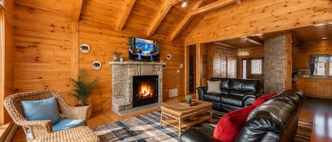 A log cabin interior with a fireplace, leather sofa, wicker chairs, and a television.