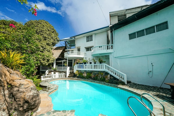 A swimming pool with a stone border, surrounded by lush greenery and a white building with a balcony.