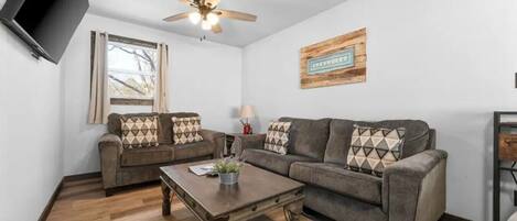 A living room with two sofas, a coffee table, a ceiling fan, a window, and a wall-mounted television.