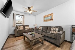 A living room with two sofas, a coffee table, a ceiling fan, a window, and a wall-mounted television. - Twin Cottages at Cardinal Hill-Sleeps 12 (Branson)