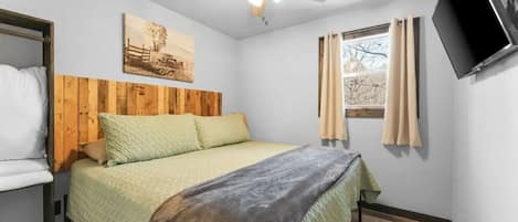 Ein Schlafzimmer mit Bett, Holz-Kopfteil, Deckenventilator, Fenster mit Vorhängen und einem an der Wand befestigten Flachbildfernseher.