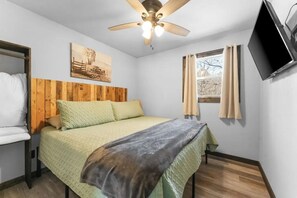 Ein Schlafzimmer mit Bett, Holz-Kopfteil, Deckenventilator, Fenster mit Vorhängen und einem an der Wand befestigten Flachbildfernseher.