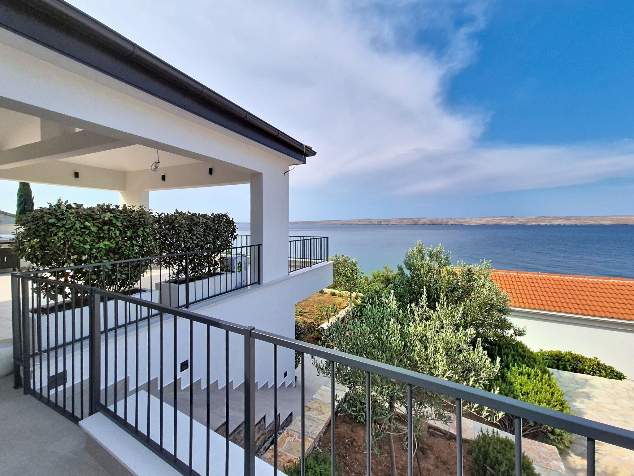 A balcony with a view of the sea, a white building, and some greenery.