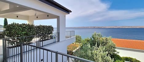 A balcony with a view of the sea, a white building, and some greenery.