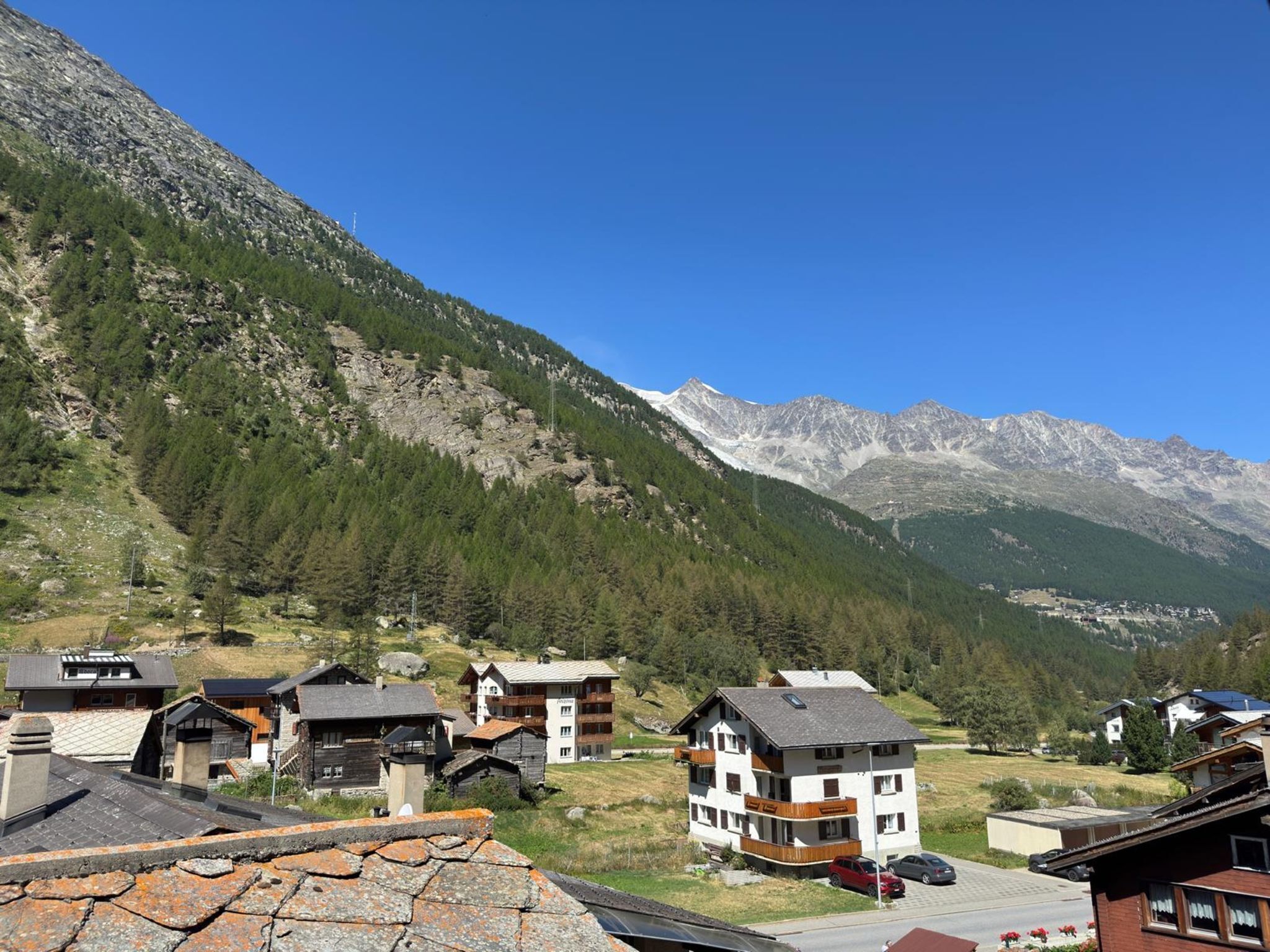 Un pueblo de montaña con casas de madera tradicionales, cielo azul despejado y una vista de montañas nevadas al fondo.