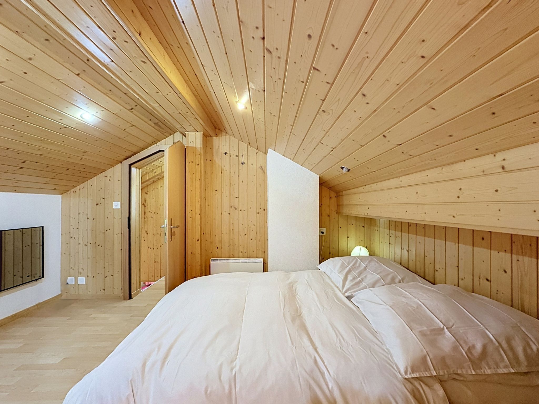 Une chambre avec un plafond en bois, un lit recouvert de draps blancs et un téléviseur fixé au mur.