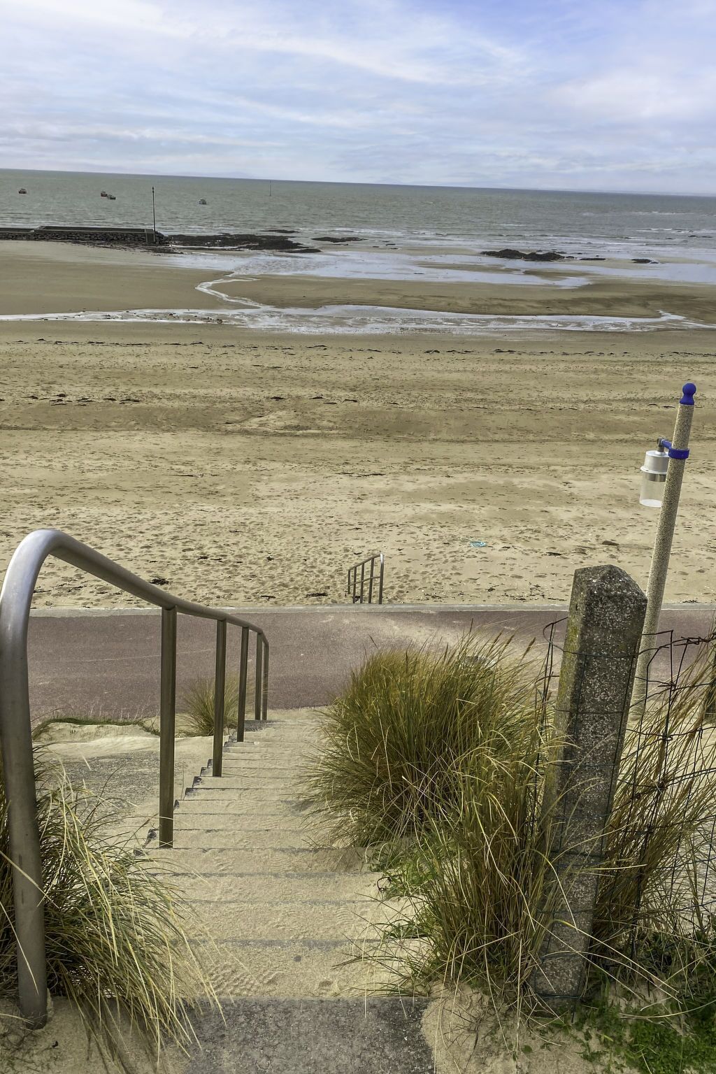 Una playa con una escalera metálica que baja hacia la orilla, un paseo marítimo de hormigón y una barandilla de metal.